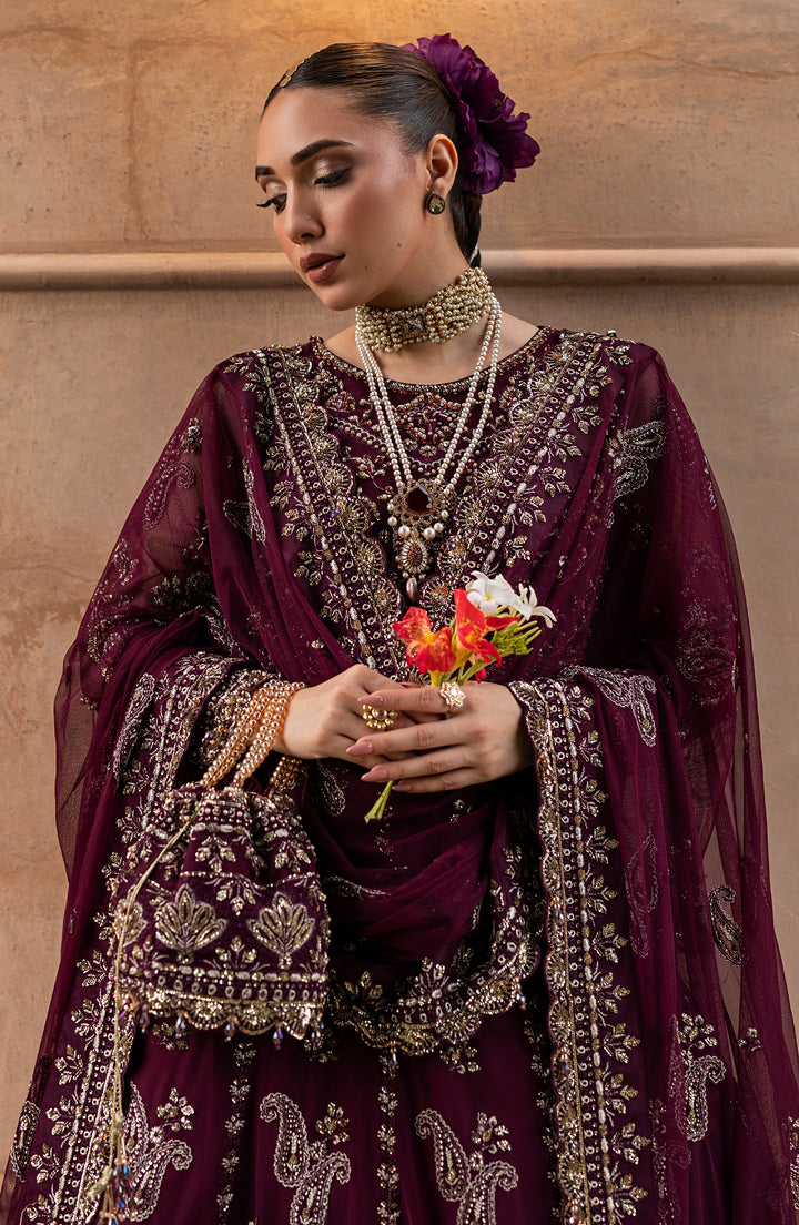 Woman in a traditional embroidered outfit holding flowers against a beige wall.